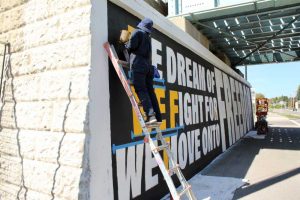 Mer painting the large yellow letters on a ladder.