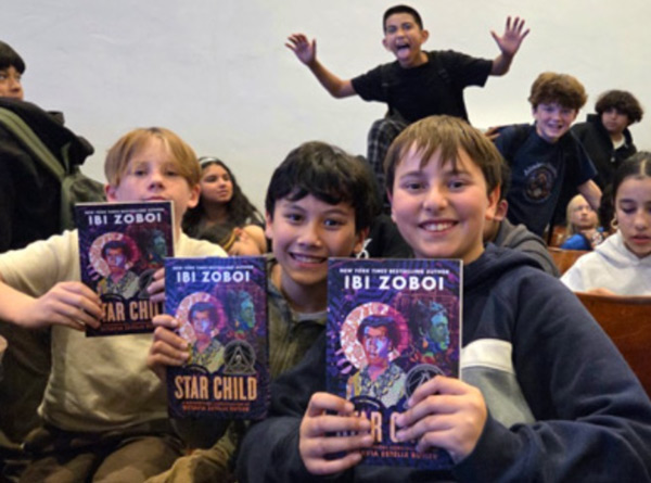 A group of smiling students inside a school auditorium proudly holding up copies of Ibi Zoboi's book, "Star Child: A Biographical Constellation of Octavia Estelle Butler," during the Octavia Fest event.